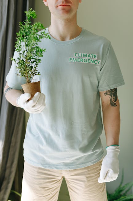 A person wearing a light grey T-shirt with the words 'Climate Emergency' printed on it, holding a small potted plant with bright green, healthy leaves. The individual is wearing white gloves, with vis