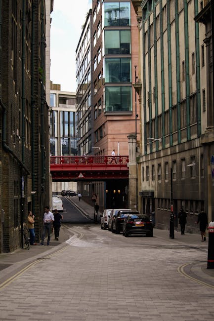 The image depicts a narrow urban street in the city, flanked by tall modern buildings with glass and stone facades. At the far end of the street, a red decorative bridge spans above, connecting the bu