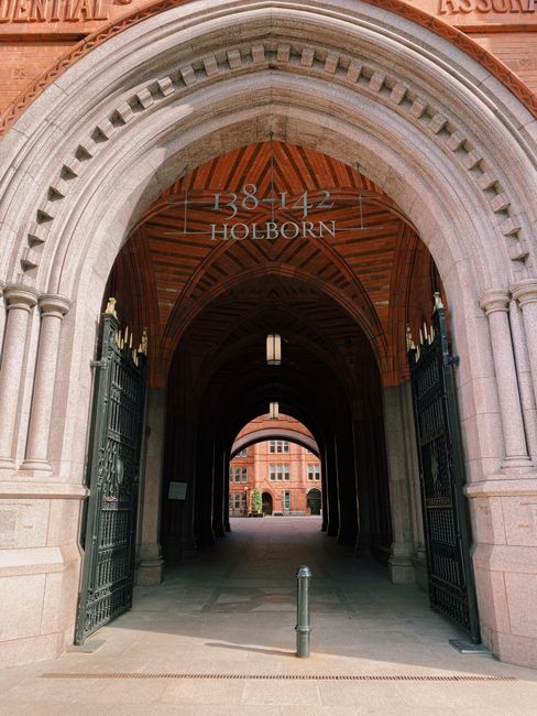 The image depicts a narrow urban street in the city, flanked by tall modern buildings with glass and stone facades. At the far end of the street, a red decorative bridge spans above, connecting the bu